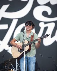 Wyatt Flores smiling while playing a guitar, wearing black sunglasses, a green striped revere shirt and blue jeans in front of a black and white backdrop.