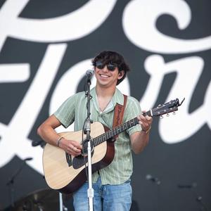 Wyatt Flores smiling while playing a guitar, wearing black sunglasses, a green striped revere shirt and blue jeans in front of a black and white backdrop.