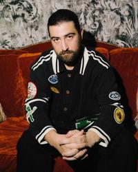 Noah Kahan sat on a sofa wearing a baseball jacket with patches 