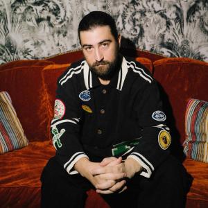 Noah Kahan sat on a sofa wearing a baseball jacket with patches 