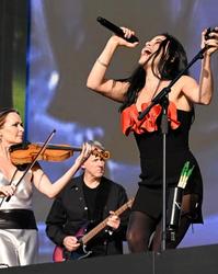 Andrea Corr sings into a microphone in a black dress with red bow while holding a microphone stand as Sharon Corr plays violin behind her on stage at BST: Hyde Park.