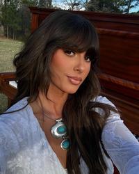 Ella Langley taking a selfie photo outside wearing a white top standing in front of a brown piano.