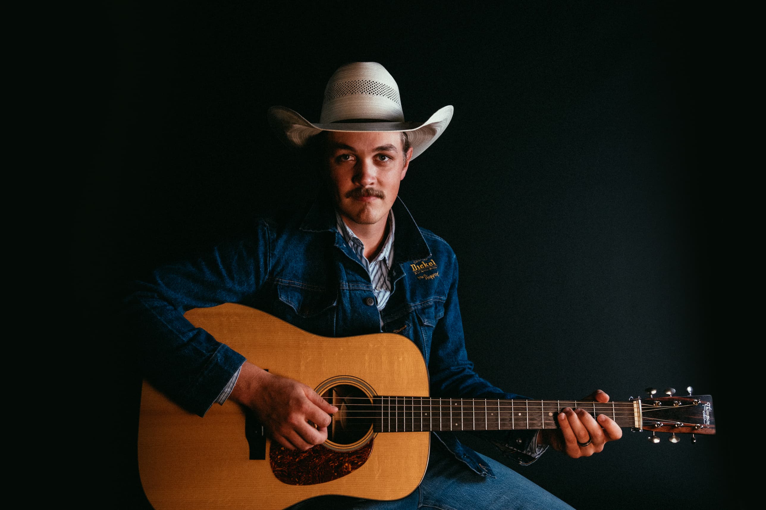 Zach Top, in front of a black background, sits and stares into the camera while wearing a denim George Dickel jacket, white cowboy hat, and blue and white striped shirt and denim jeans, while holding a guitar.