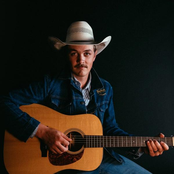 Zach Top, in front of a black background, sits and stares into the camera while wearing a denim George Dickel jacket, white cowboy hat, and blue and white striped shirt and denim jeans, while holding a guitar.