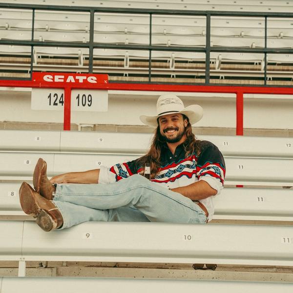 Ian Munsick smiling in a cowboy hat while sat in the empty stands with his legs crossed over the front row