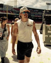 Tucker Wetmore in a sleeveless tee and black shorts, sunglasses and trucker cap, walking through a stadium.