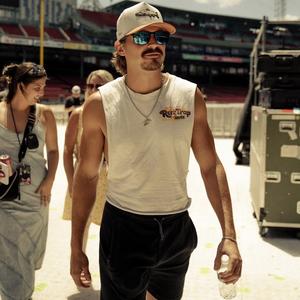 Tucker Wetmore in a sleeveless tee and black shorts, sunglasses and trucker cap, walking through a stadium.