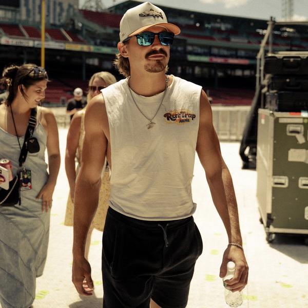 Tucker Wetmore in a sleeveless tee and black shorts, sunglasses and trucker cap, walking through a stadium.
