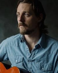 Sturgill Simpson staring to the left wearing a blue denim shirt, holding a guitar while sat in front of a black background.