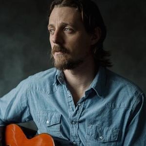 Sturgill Simpson staring to the left wearing a blue denim shirt, holding a guitar while sat in front of a black background.