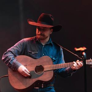 Colter Wall in front of a blue neon sign on stage at Two Step Inn, wearing a black cowboy hat, denim jacket and playing a guitar.