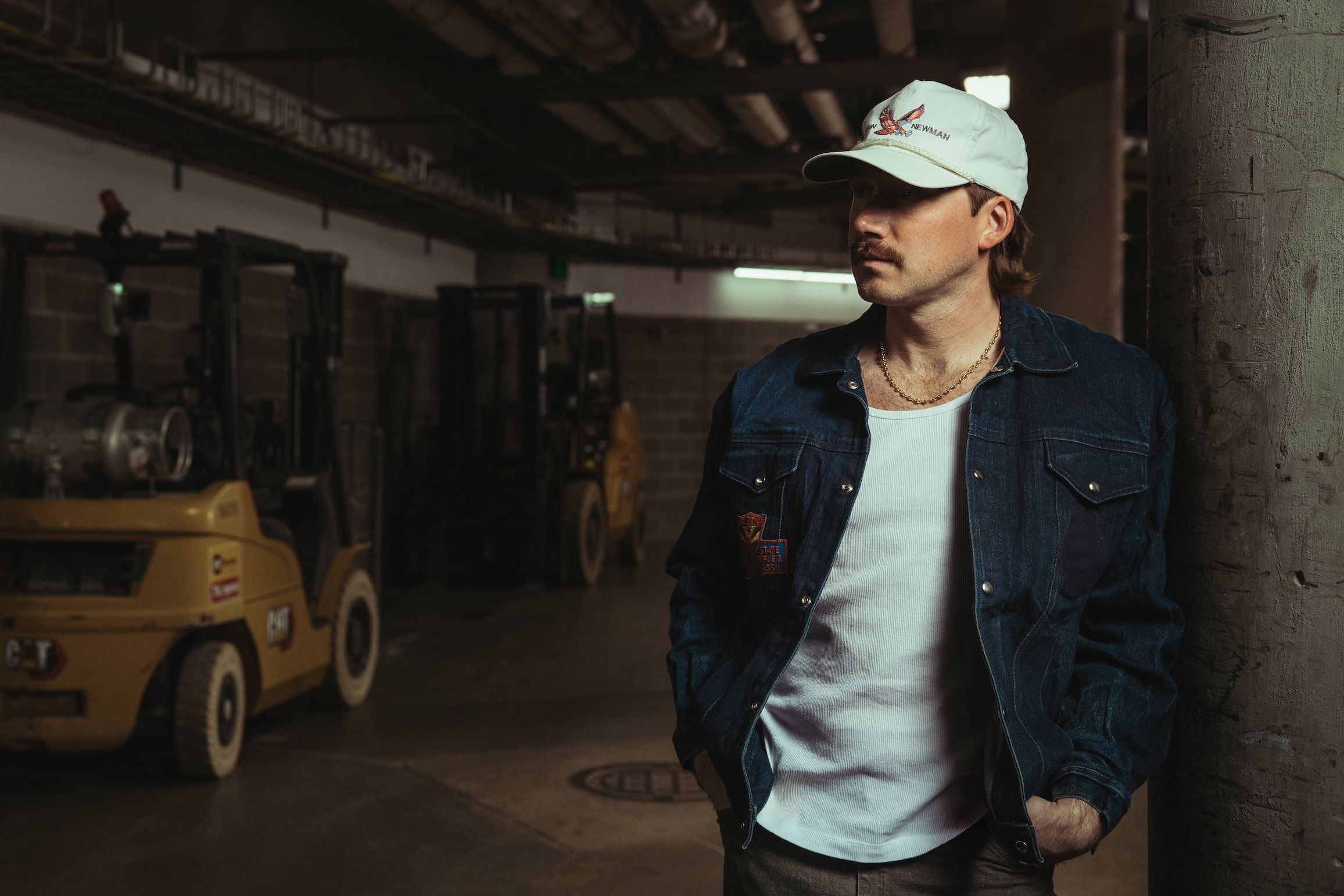 Morgan Wallen wearing a white vest, denim shirt jacket and white-grey cap, standing in a stadium backstage with heavy duty vehicles next to him.