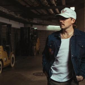 Morgan Wallen wearing a white vest, denim shirt jacket and white-grey cap, standing in a stadium backstage with heavy duty vehicles next to him.