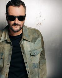 Eric Church in a camo jacket and black t-shirt wearing black sunglasses while infront of a white and black wall.