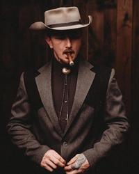 Benjamin Tod dress in full suit jacket and bolo tie, grey cowboy hat while smoking a cigar.