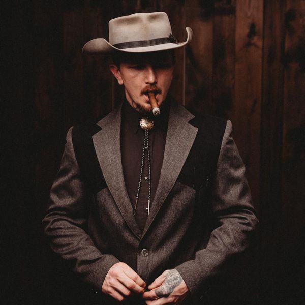 Benjamin Tod dress in full suit jacket and bolo tie, grey cowboy hat while smoking a cigar.