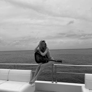 A black & white photo of Megan Moroney holding a guitar while sat on a barrier of the edge of a boat.