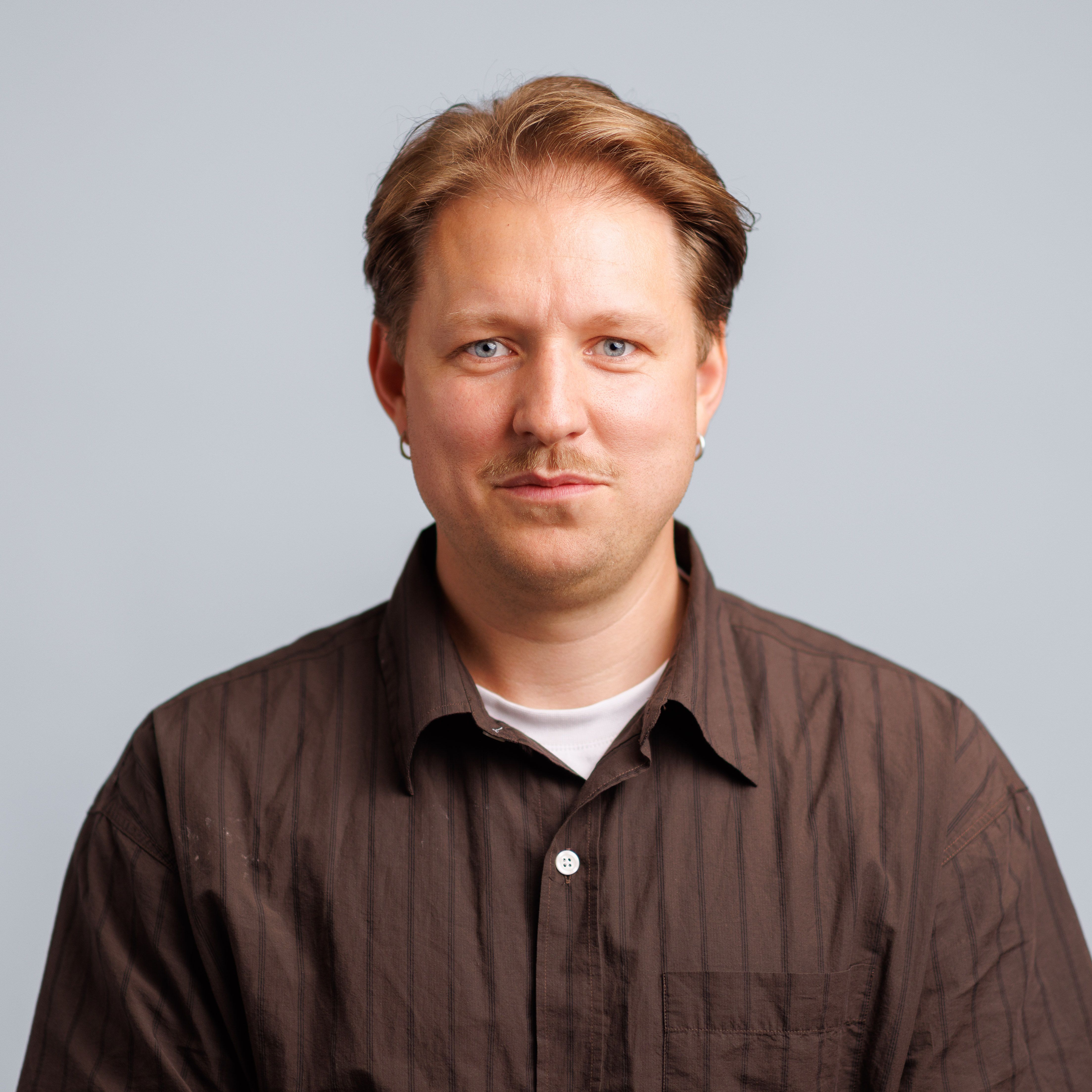 A man with light brown hair, blue eyes, a mustache, and earrings, wearing a brown striped shirt, looking forward.