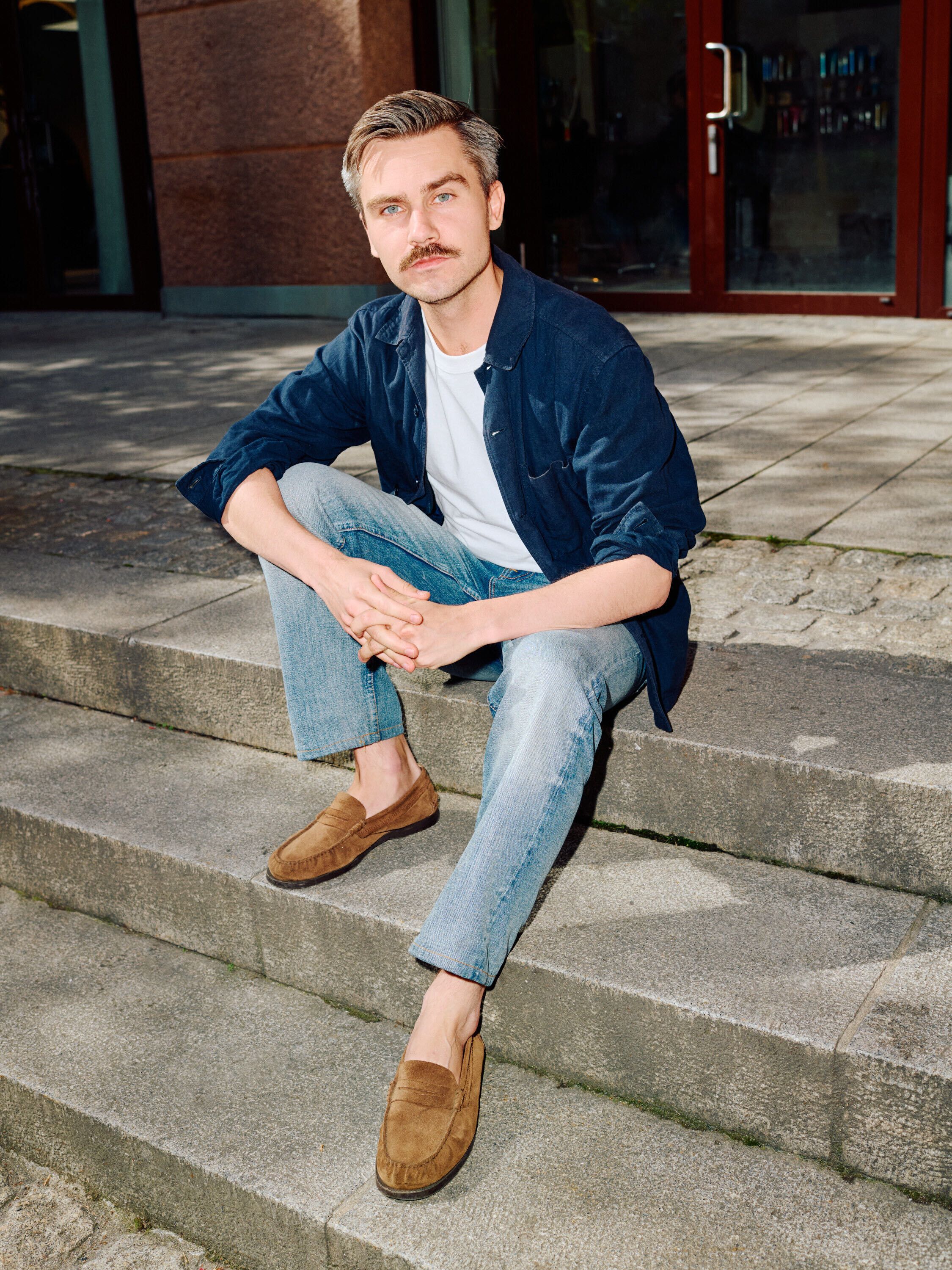 A man with a mustache and brown hair sits on stone steps, wearing a blue shirt, jeans, and brown loafers.