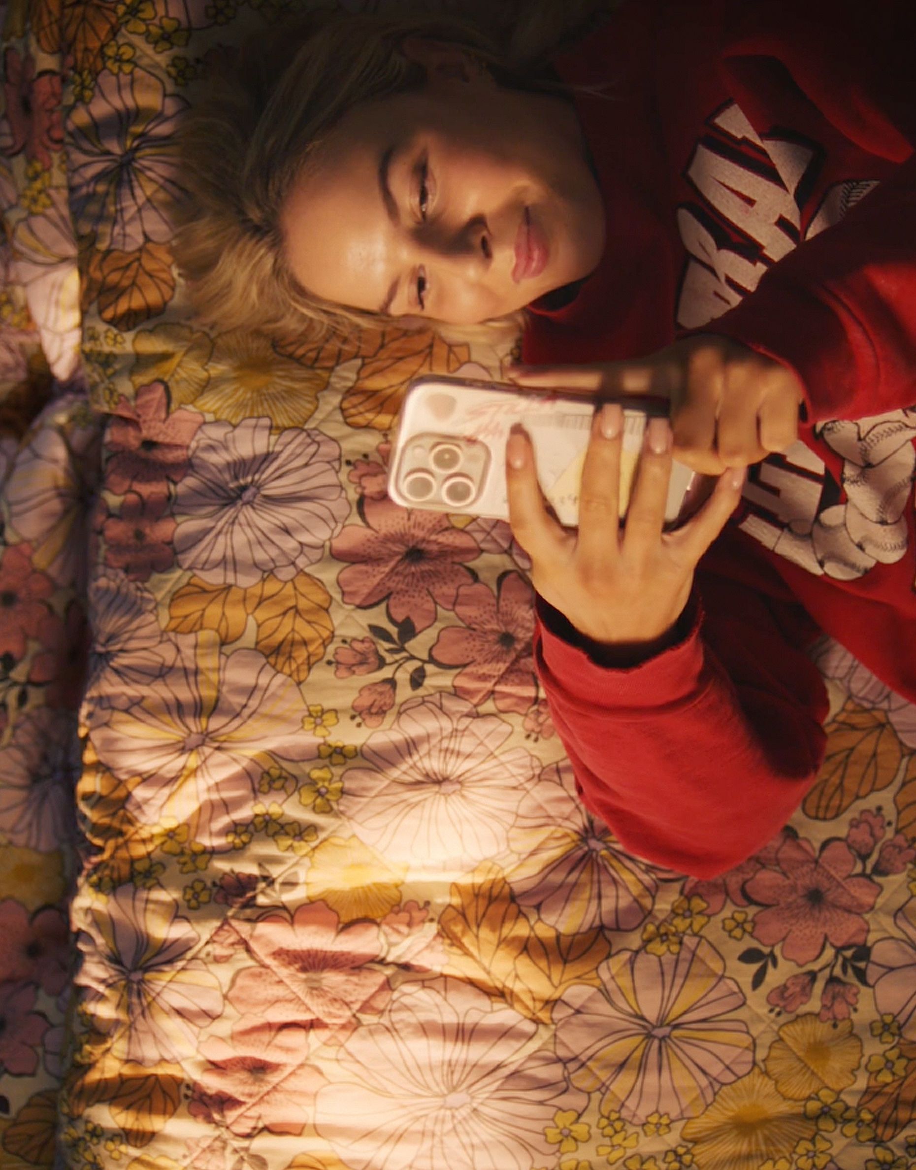 A top-down phone-in-hand shot of a young woman in a red sweatshirt lying on a floral bedspread, smiling at her phone, with a second person visible at the edge of frame.