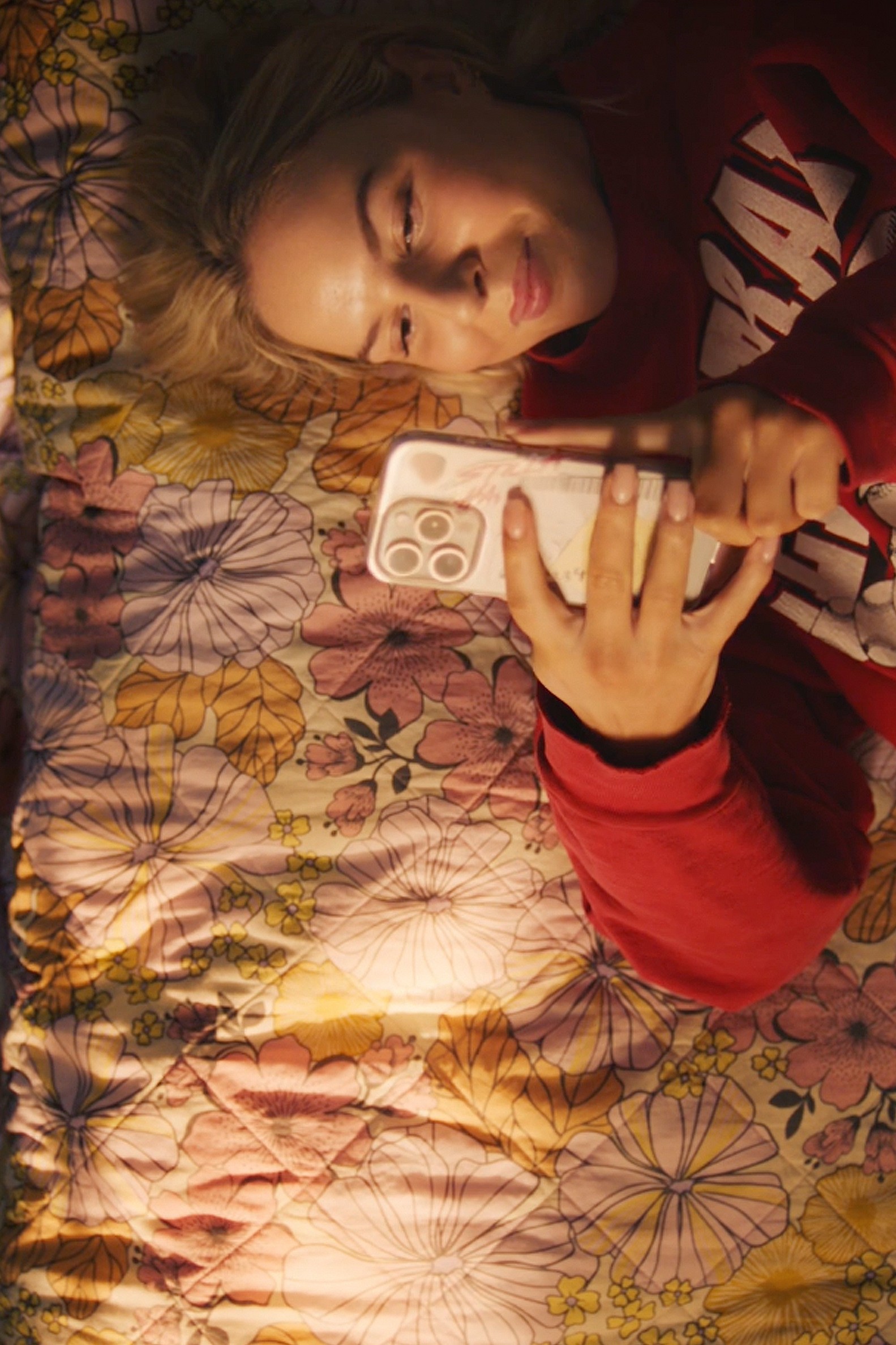 A top-down phone-in-hand shot of a young woman in a red sweatshirt lying on a floral bedspread, smiling at her phone, with a second person visible at the edge of frame.