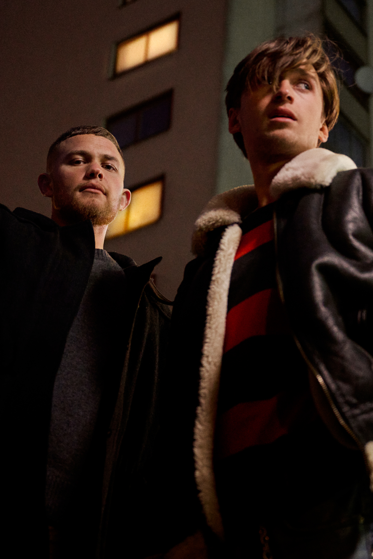 A production still of two young men on a bed, one lying down using a phone and the other sitting up looking at him, captioned "A series about drunkenness, betrayal & love" in orange type.