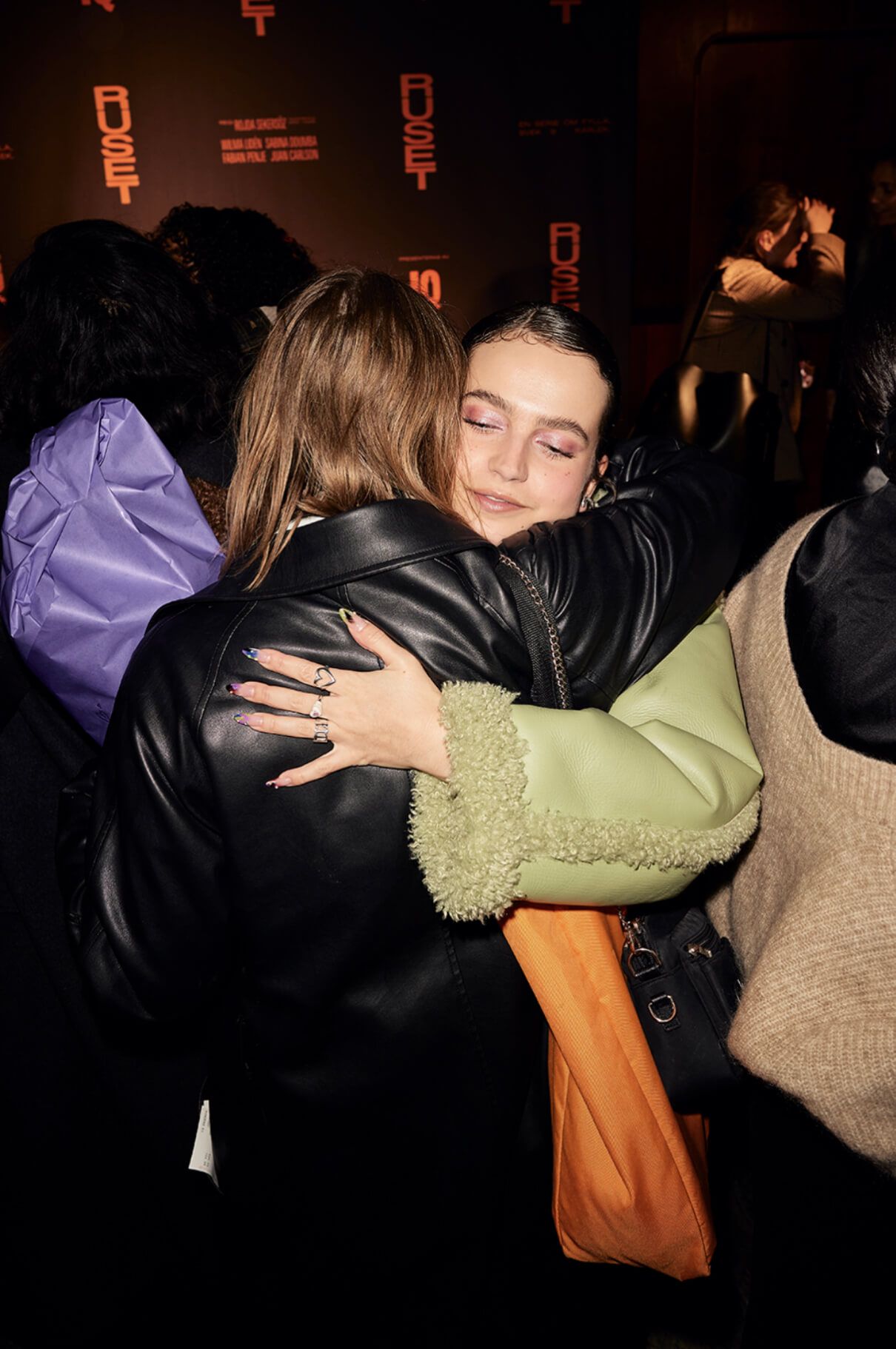 Two young women embracing in a crowd at the Ruset premiere, in front of a RUSET step-and-repeat backdrop.