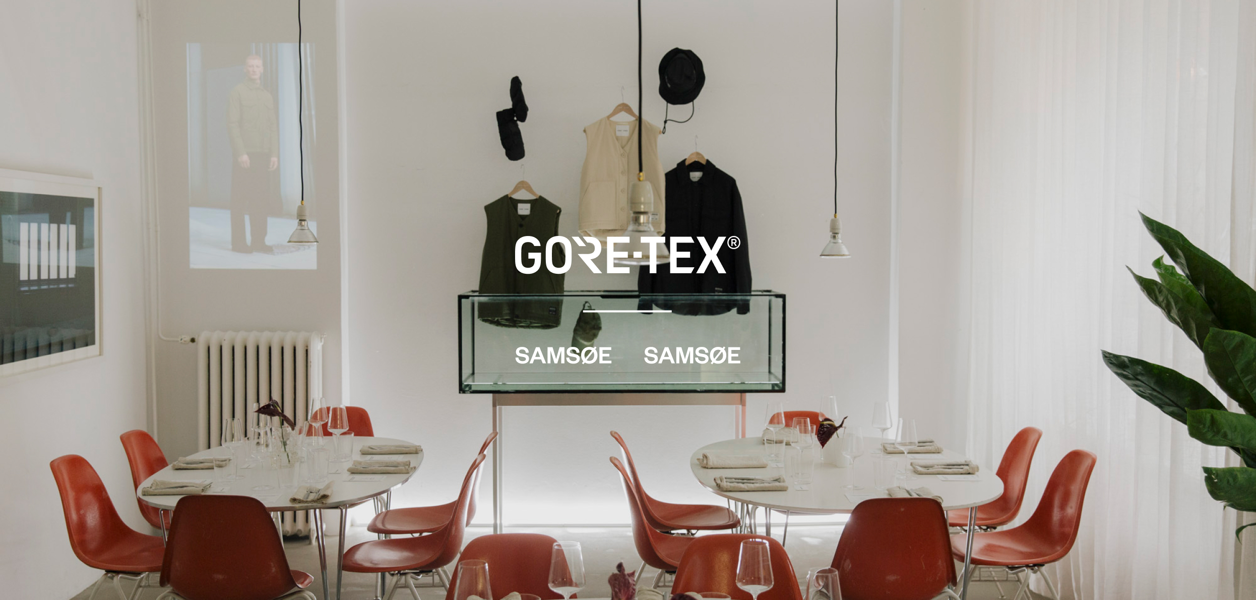 A minimalist room featuring dining tables, modern orange chairs, and a central display of Gore-Tex and Samsøe Samsøe clothing, with a projected image of a model.