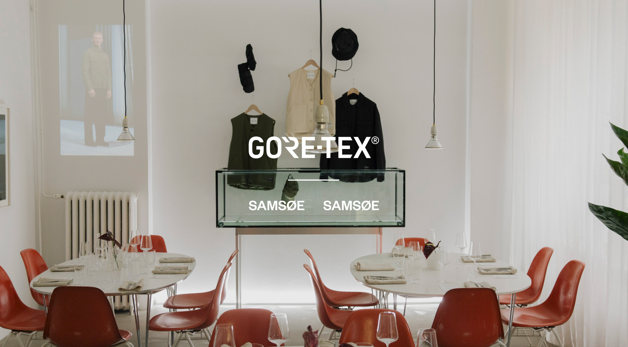 A minimalist room featuring dining tables, modern orange chairs, and a central display of Gore-Tex and Samsøe Samsøe clothing, with a projected image of a model.