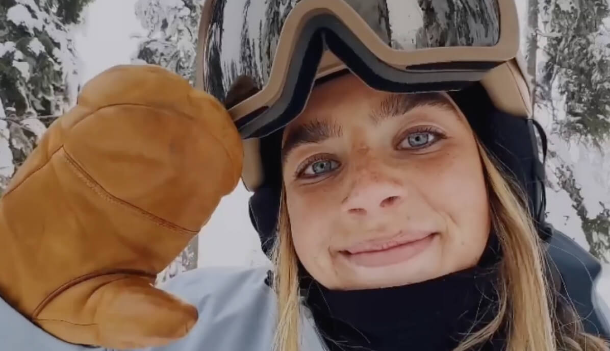A smiling skier holding up a mitten to the camera, goggles pushed up, forest behind.