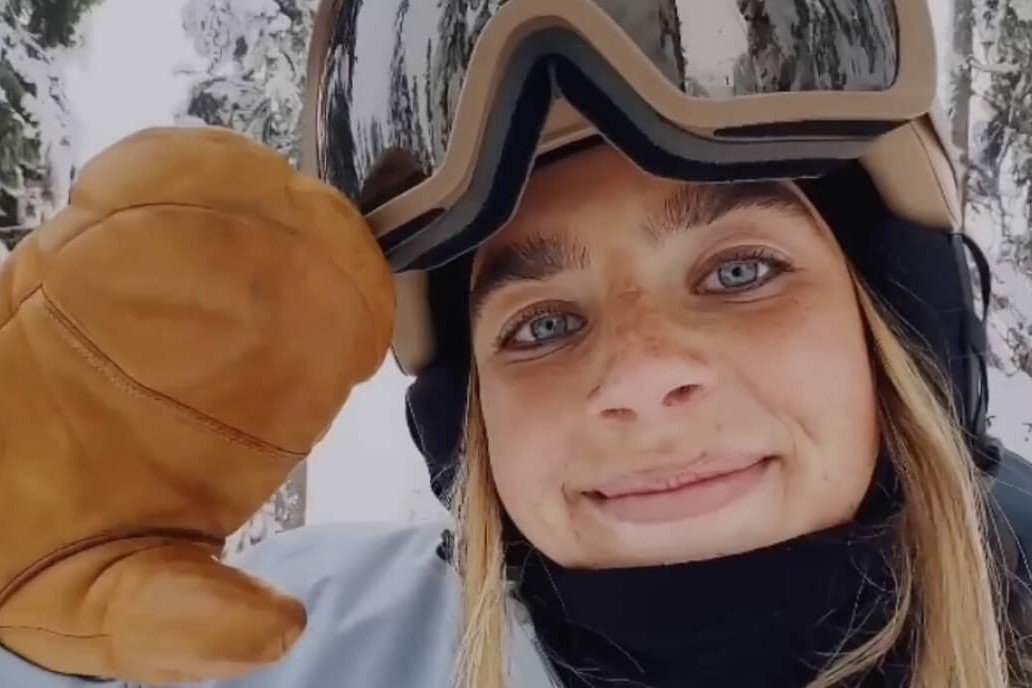 A smiling skier holding up a mitten to the camera, goggles pushed up, forest behind.