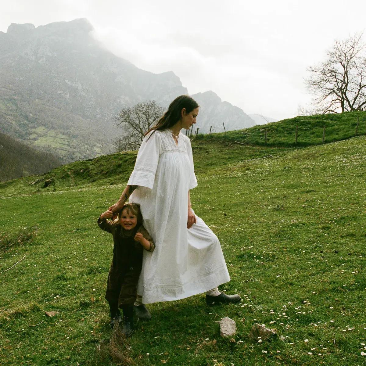 A woman in a white dress holds the hand of a smiling child on a grassy hill dotted with flowers, with misty mountains in the distance.