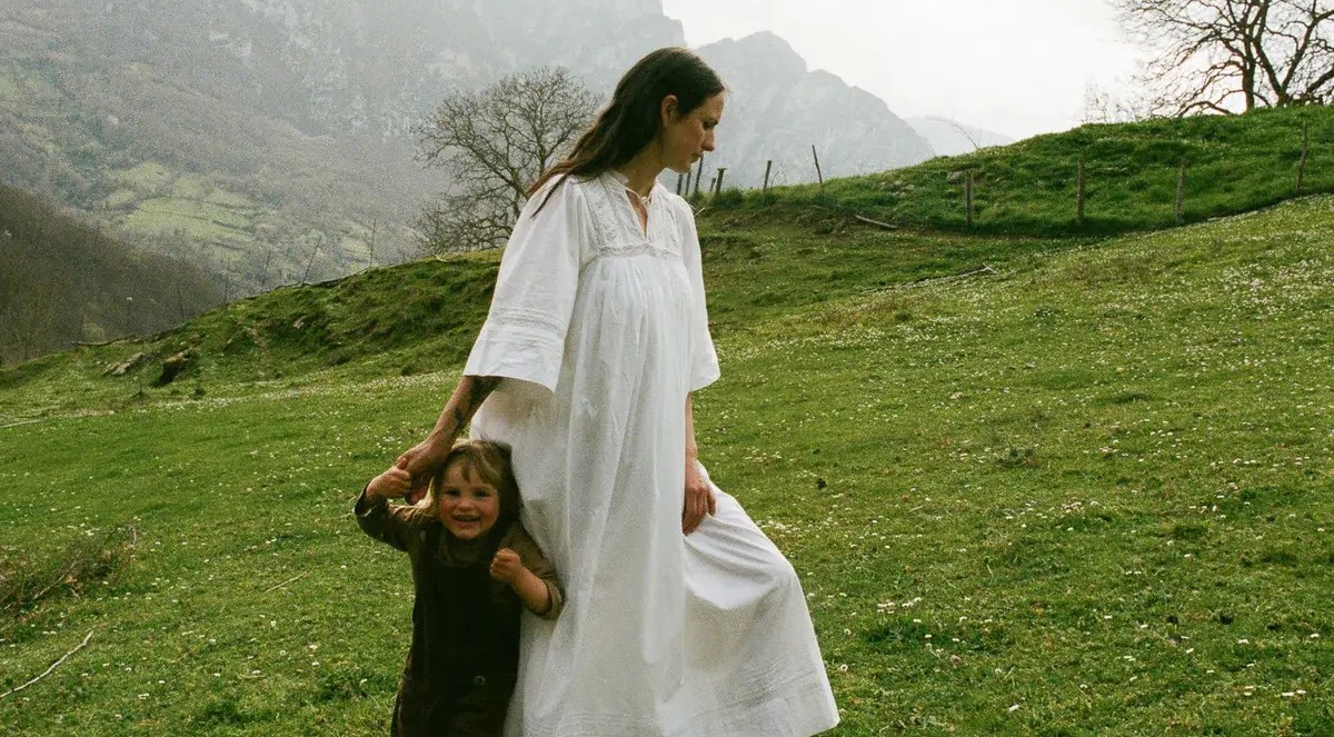 A woman in a white dress holds the hand of a smiling child on a grassy hill dotted with flowers, with misty mountains in the distance.