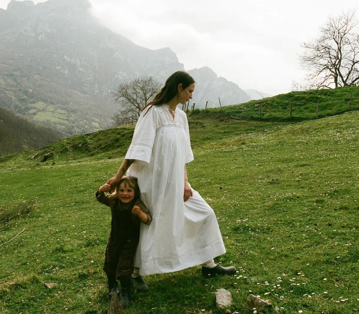A woman in a white dress holds the hand of a smiling child on a grassy hill dotted with flowers, with misty mountains in the distance.