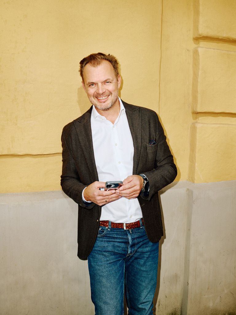 A smiling man in a blazer and jeans holds a smartphone against a yellow wall.