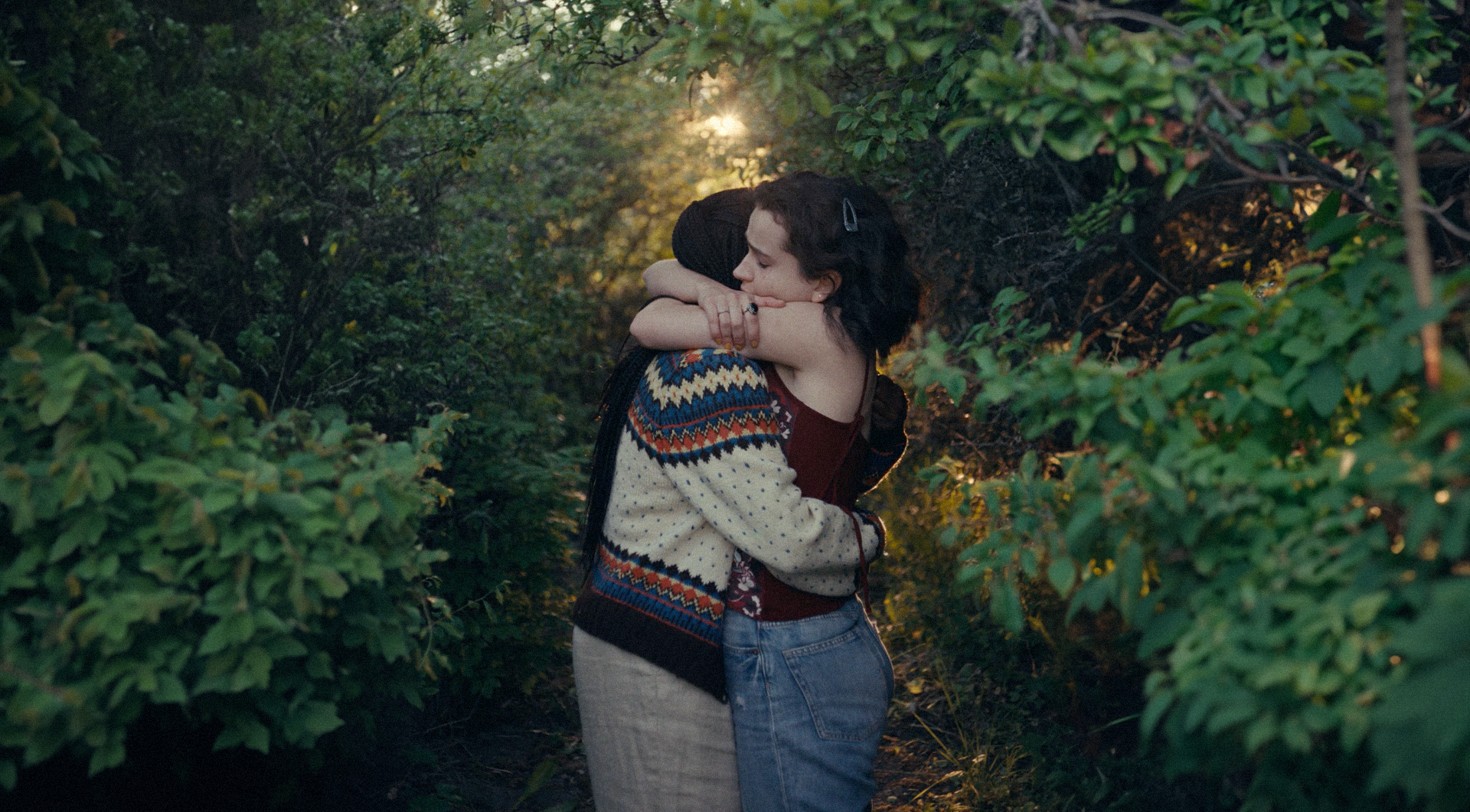 Two people embracing in a leafy outdoor setting with sunlight.