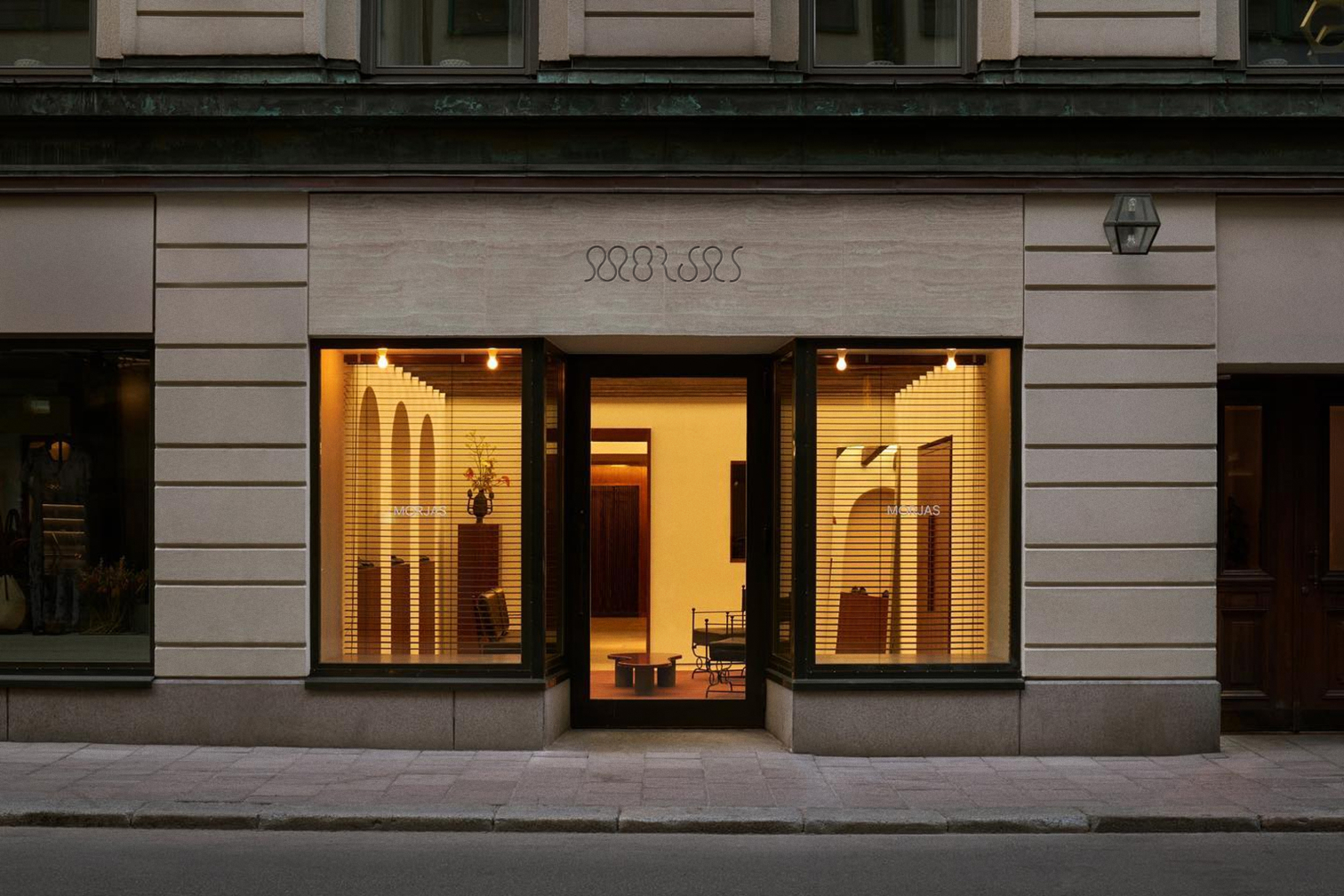 Storefront of MORJAS with illuminated windows, partially obscured by horizontal blinds, and an open entrance.