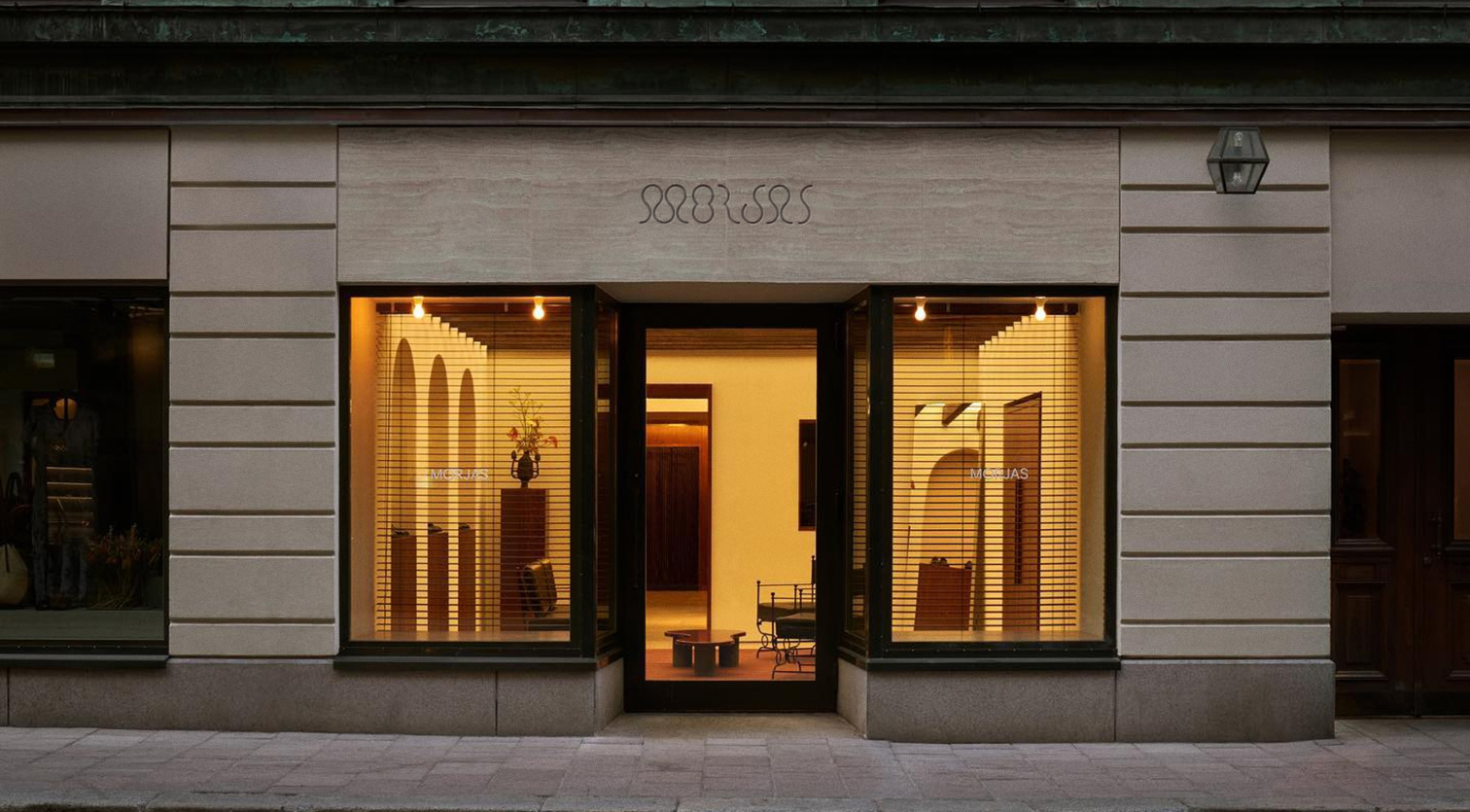 Storefront of MORJAS with illuminated windows, partially obscured by horizontal blinds, and an open entrance.