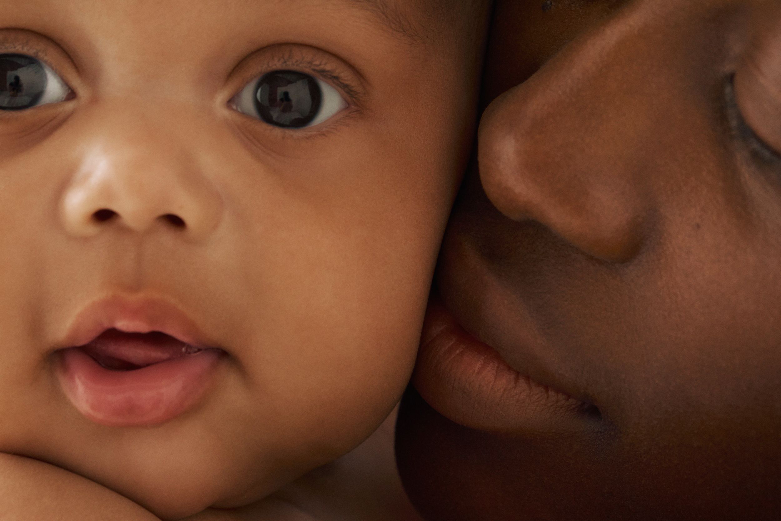 Close-up of a brown-skinned baby's face next to an adult's face, cheek-to-cheek.