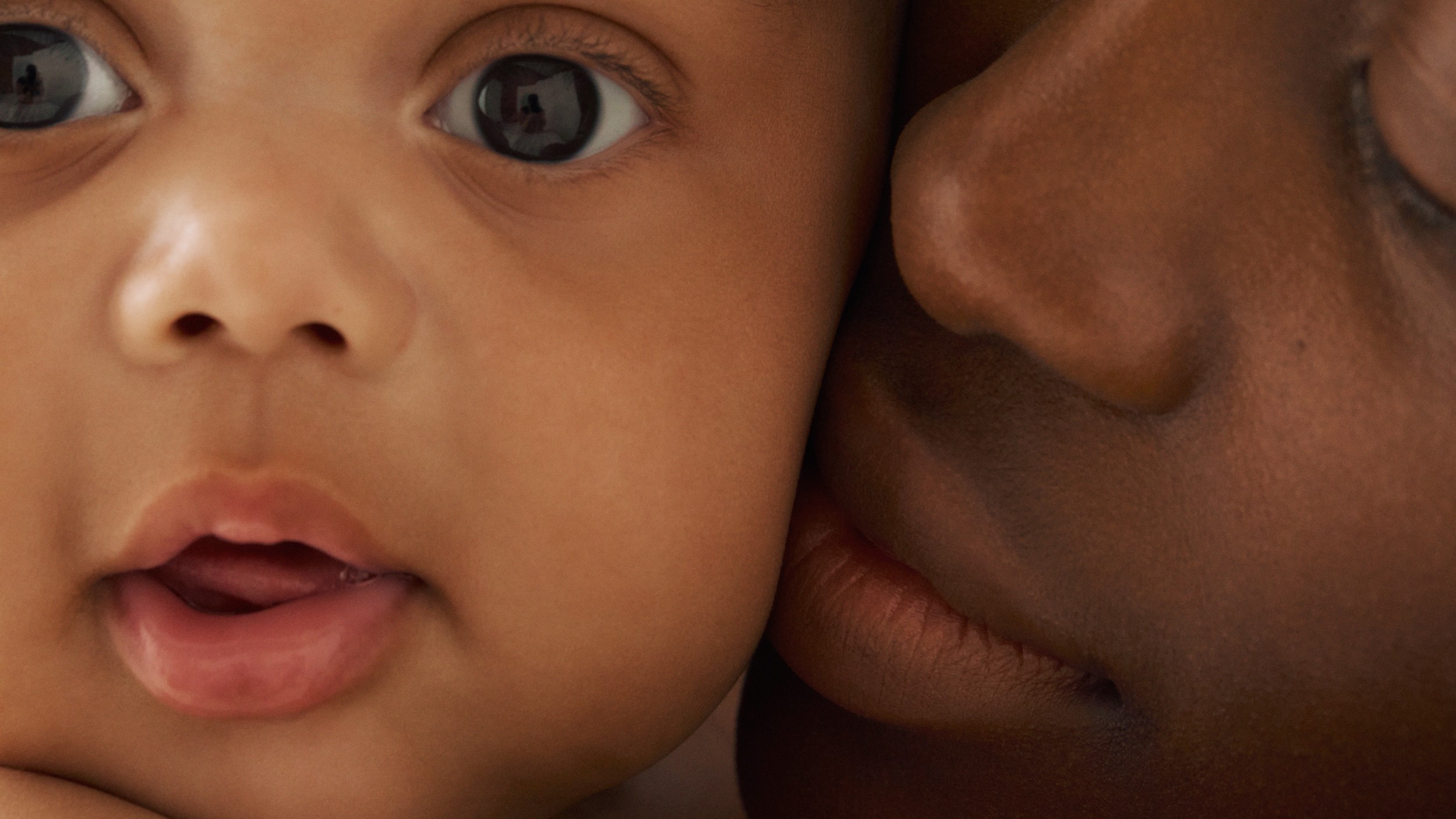 Close-up of a brown-skinned baby's face next to an adult's face, cheek-to-cheek.
