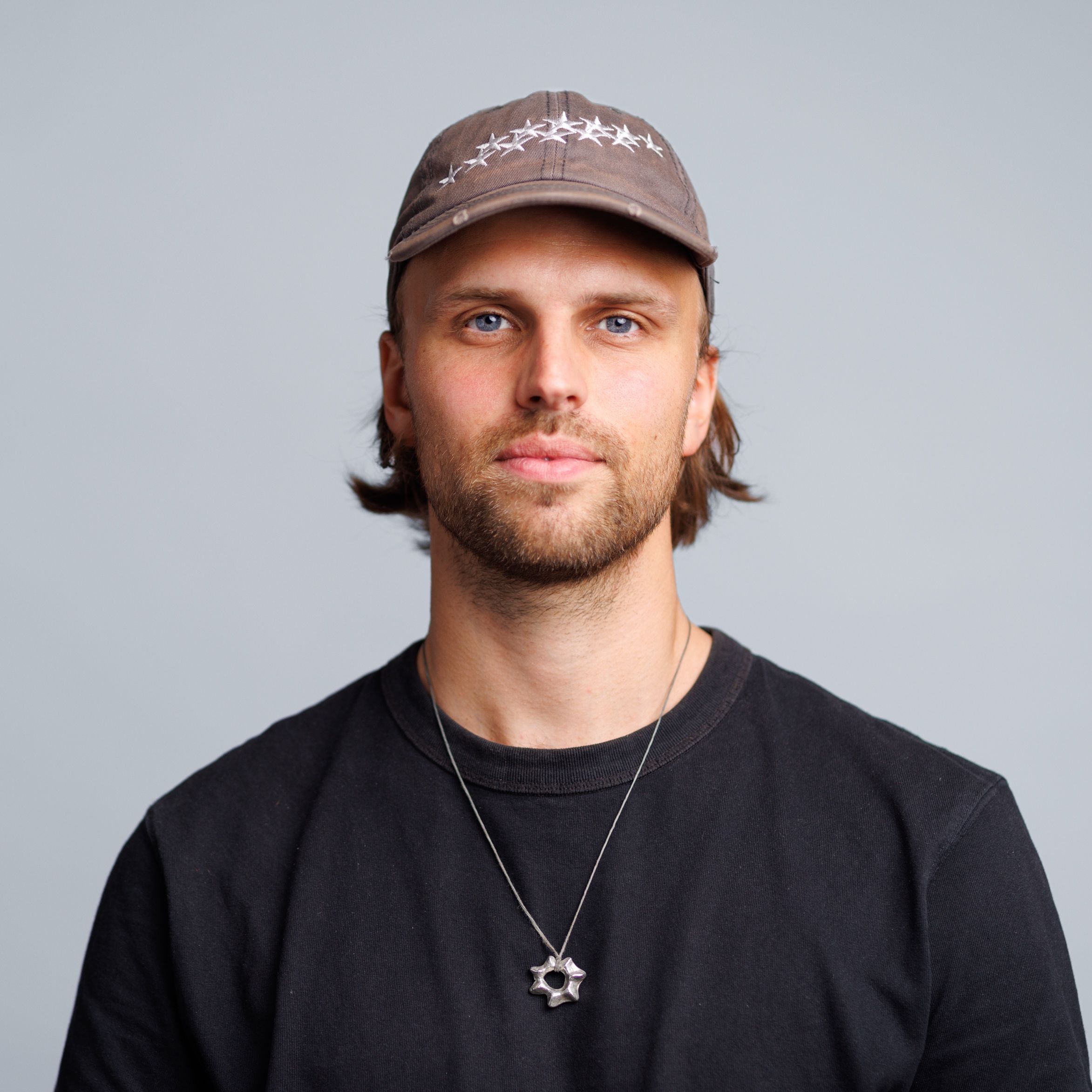 Headshot of a man with a beard, wearing a brown cap adorned with white stars and a silver multi-pointed pendant.