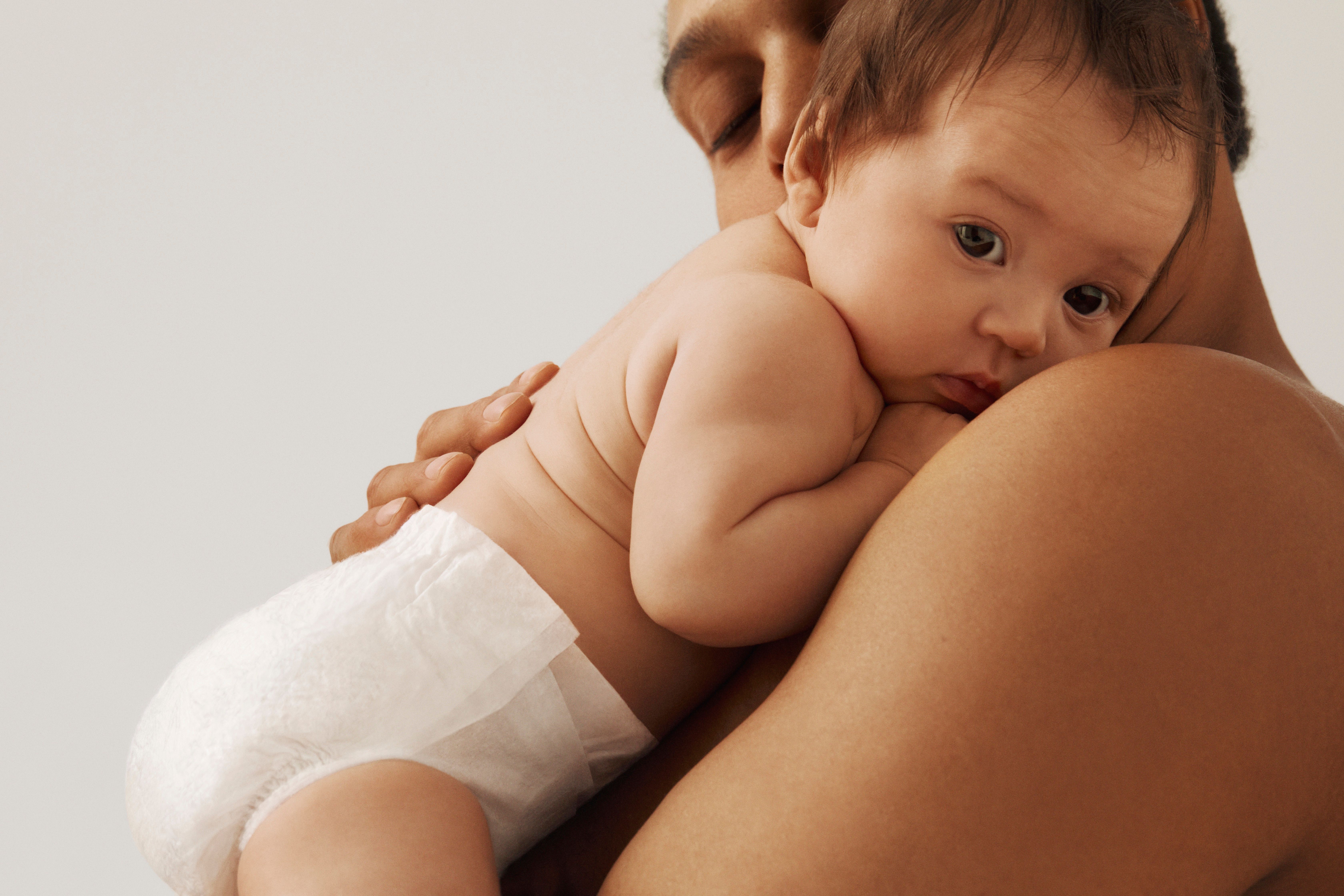 A baby in a white diaper rests on an adult's shoulder, looking at the viewer.