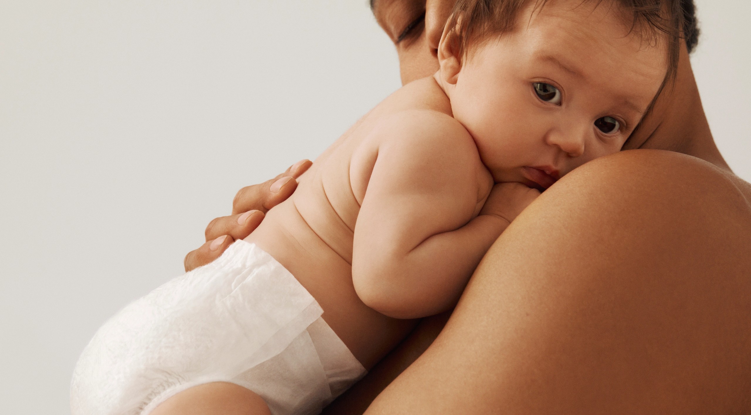 A baby in a white diaper rests on an adult's shoulder, looking at the viewer.