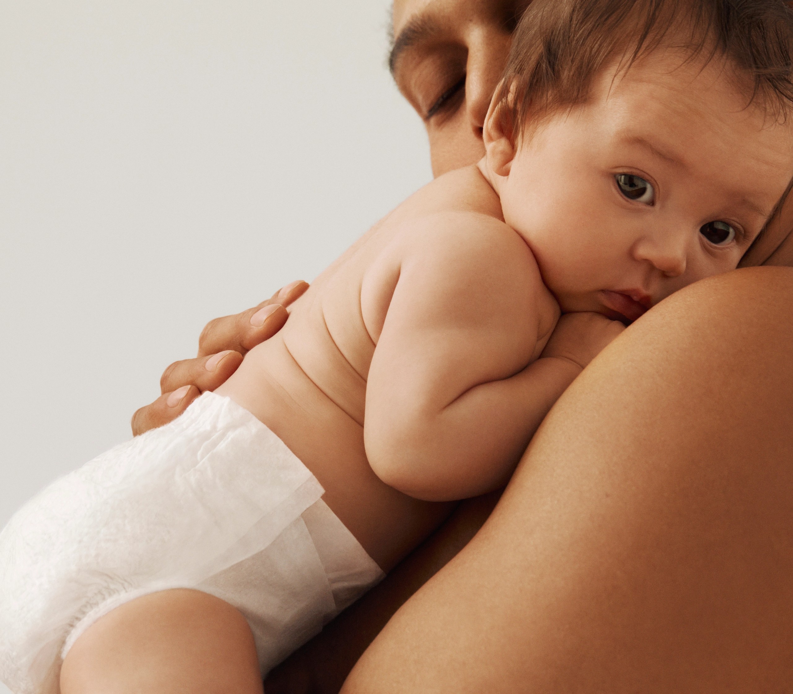 A baby in a white diaper rests on an adult's shoulder, looking at the viewer.