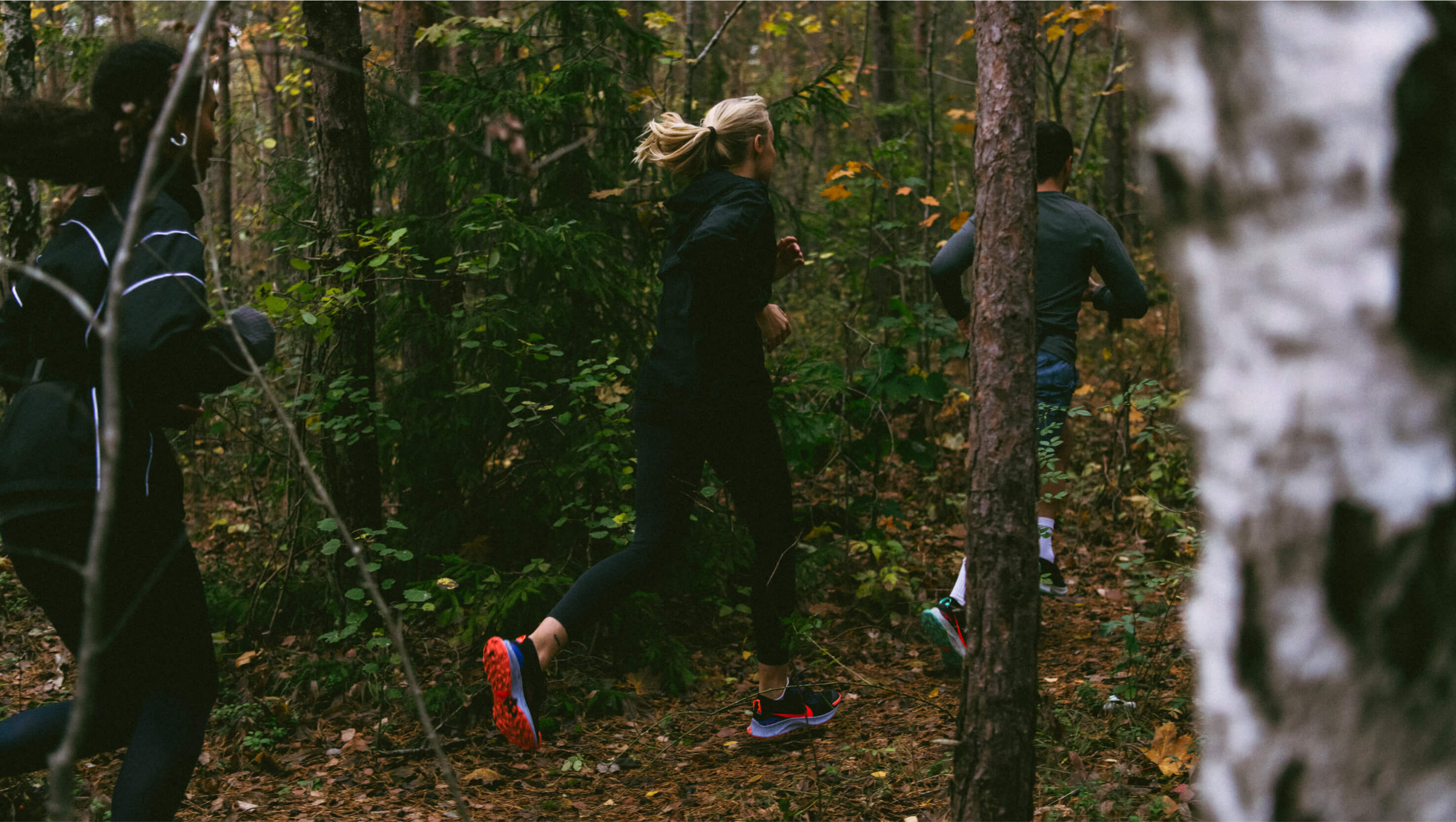 A group of runners in dark kit, one pair of orange and blue Nike trail shoes visible, moving through a forest trail in low light among autumn leaves.