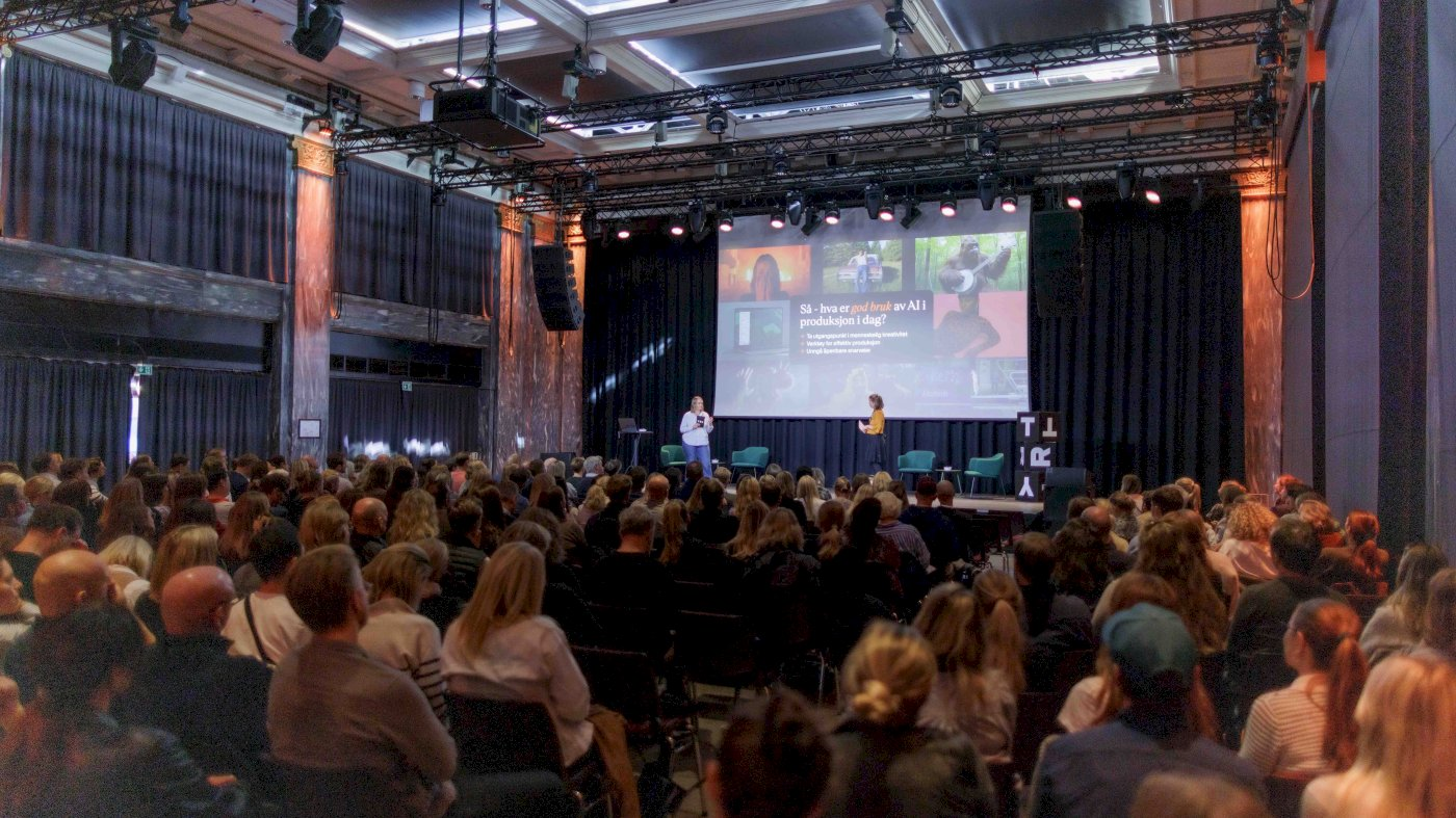 A conference hall filled with attendees watching two speakers on a stage with a presentation screen.