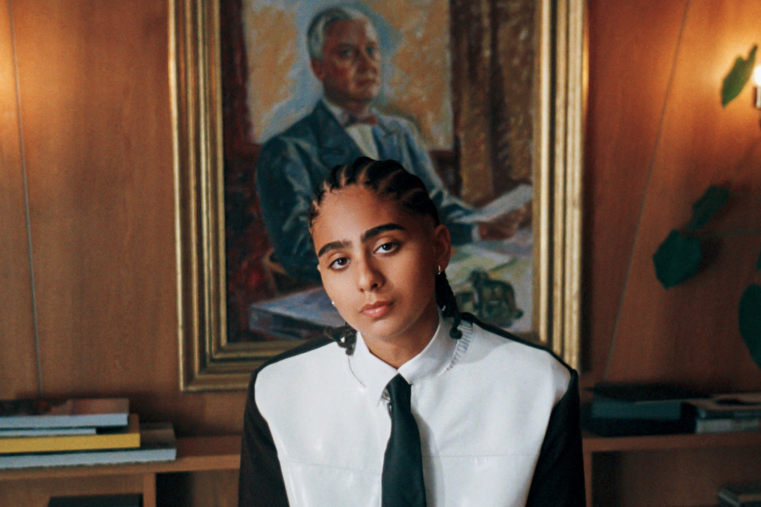 A person with braided hair and a tie looks forward, a framed portrait of an older man behind them.