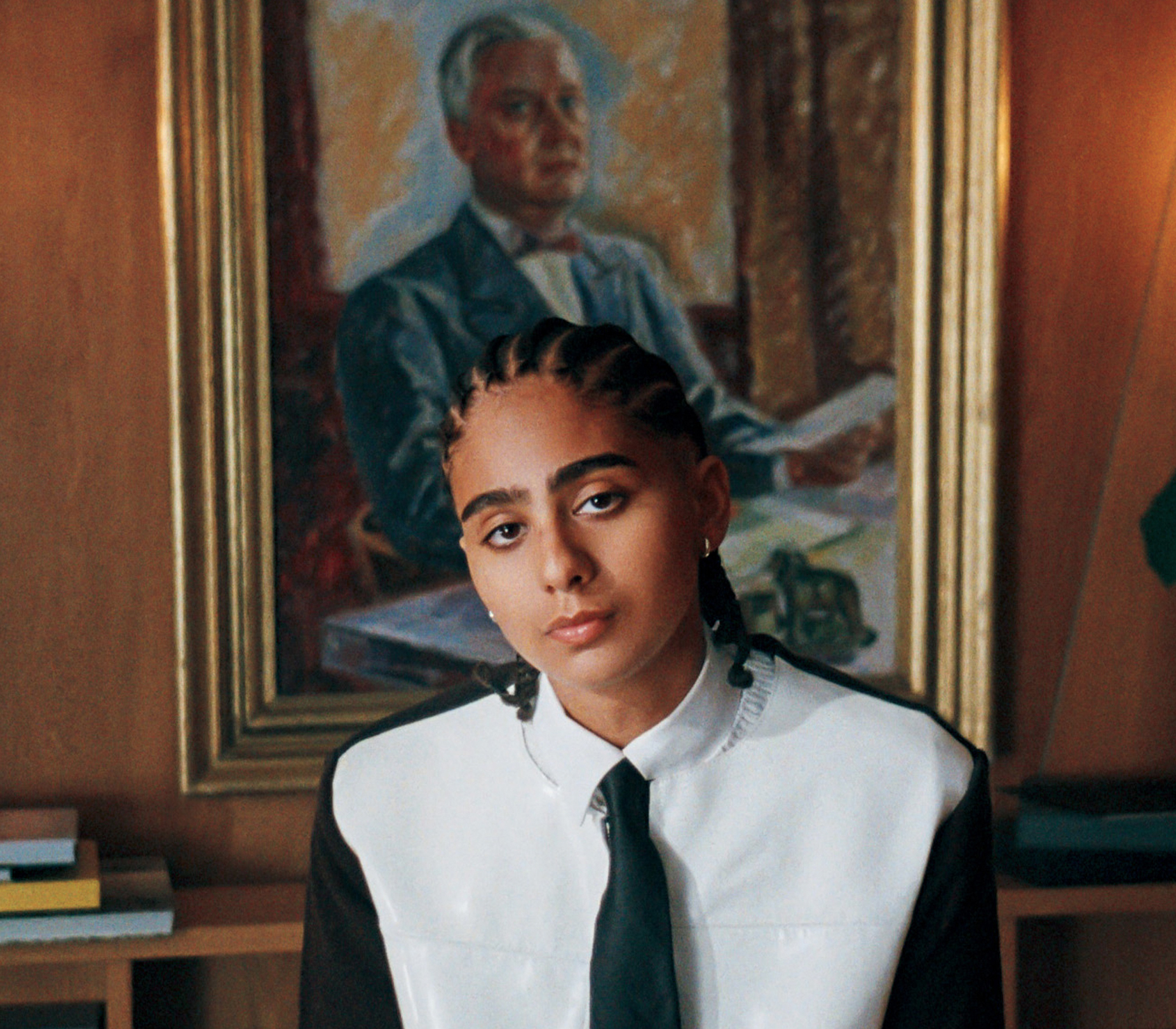 A person with braided hair and a tie looks forward, a framed portrait of an older man behind them.