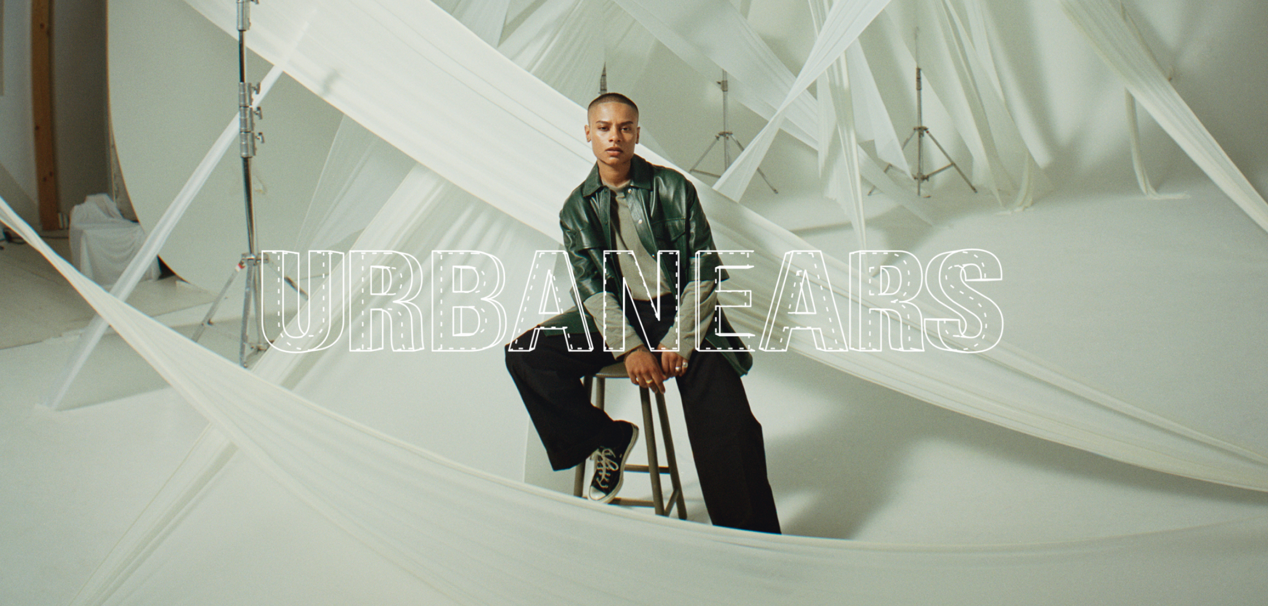 A person with a shaved head sits on a stool wearing a green jacket and black pants in a white studio draped with fabric. Text: URBANEARS.