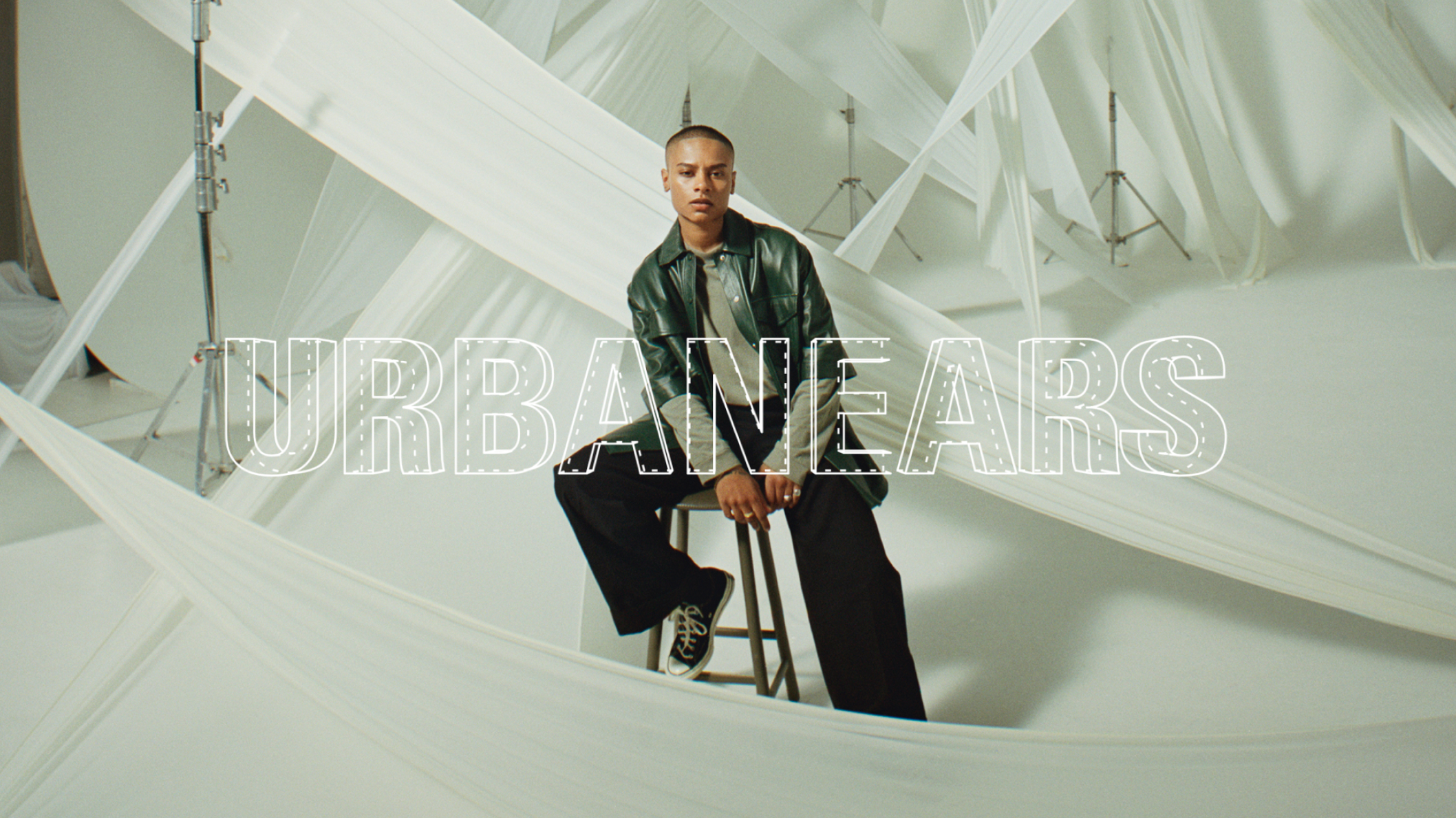 A person with a shaved head sits on a stool wearing a green jacket and black pants in a white studio draped with fabric. Text: URBANEARS.